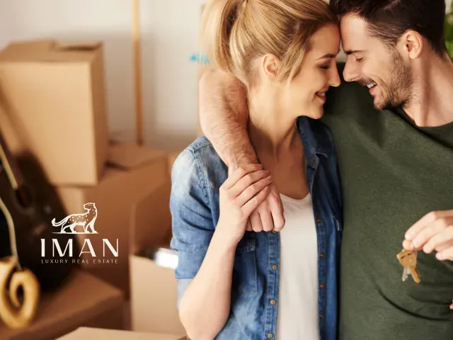 Couple embraces, holding keys, smiling near moving boxes; interior setting, logo in corner.