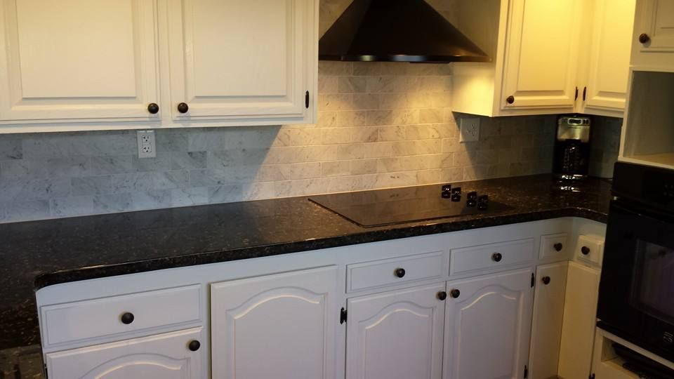 White kitchen cabinets with dark countertops, a black stove hood, and a light-colored tiled backsplash.