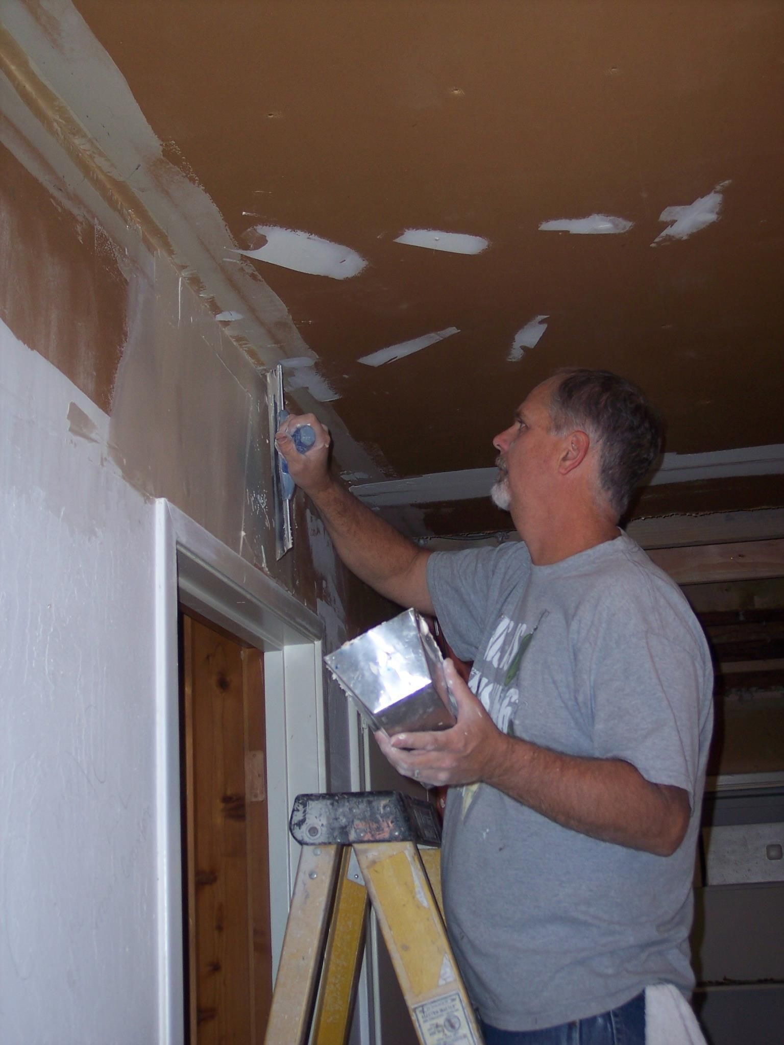 A person on a stepladder uses a taping knife to apply joint compound to the corner of a brown wall and white ceiling.