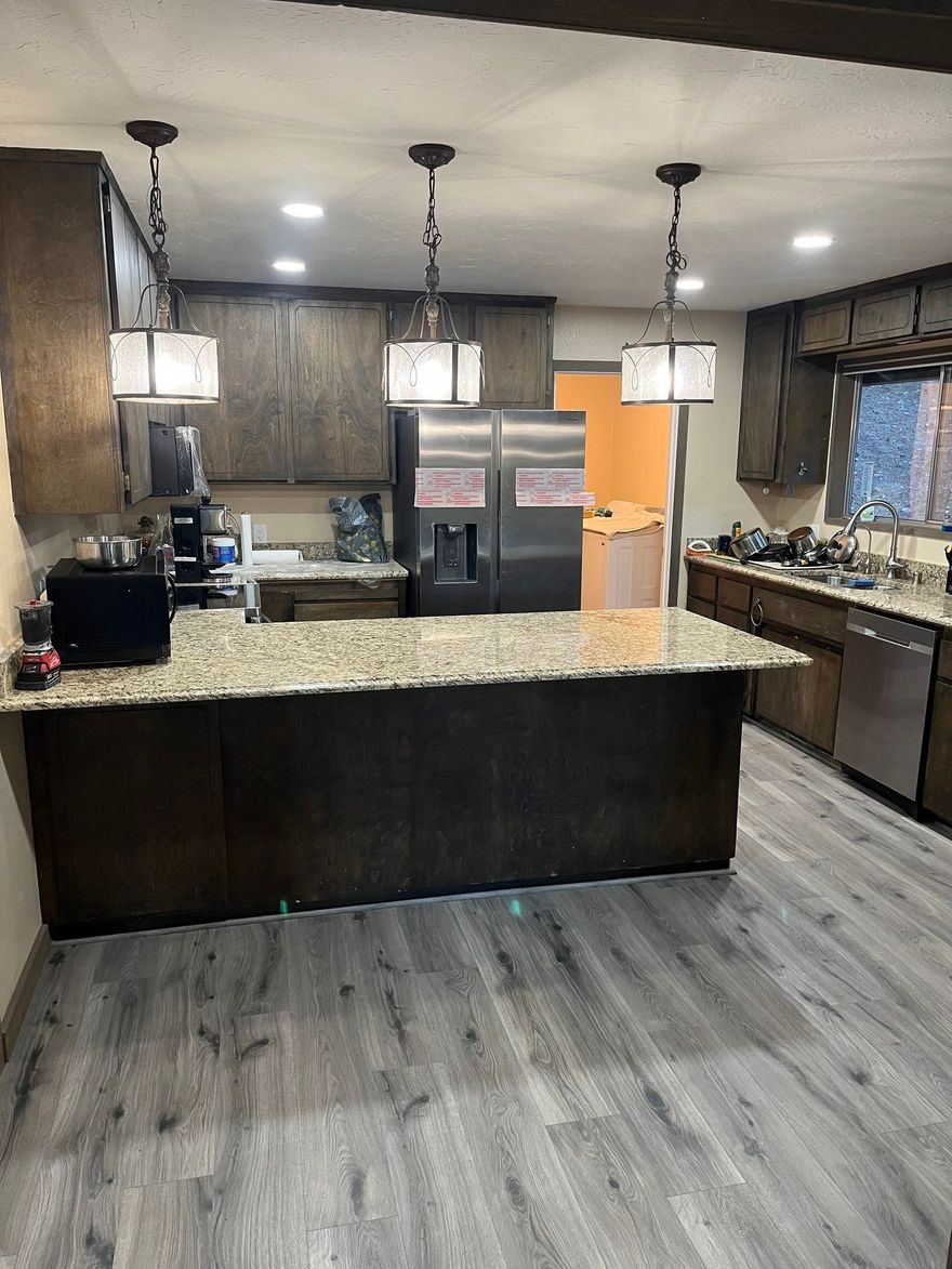 Kitchen featuring dark wood cabinets, granite countertops, a large island with three pendant lights, and gray flooring.