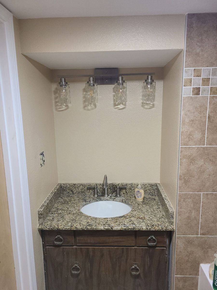 A bathroom vanity with a granite countertop, sink, and a four-light mason jar fixture mounted in a beige alcove.