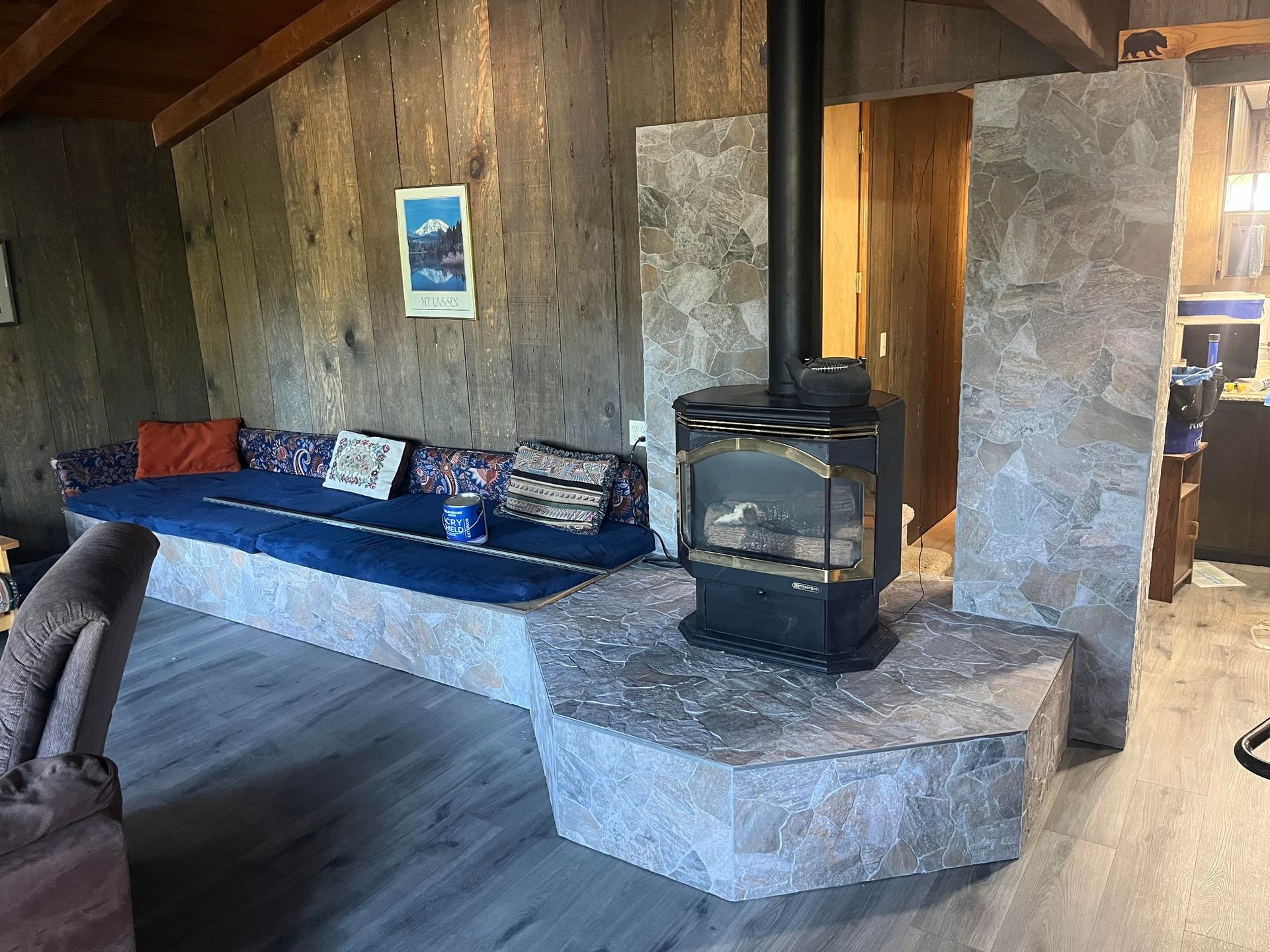 A wood-burning stove sits on a stone hearth next to a built-in bench with blue cushions against a wood-paneled wall.