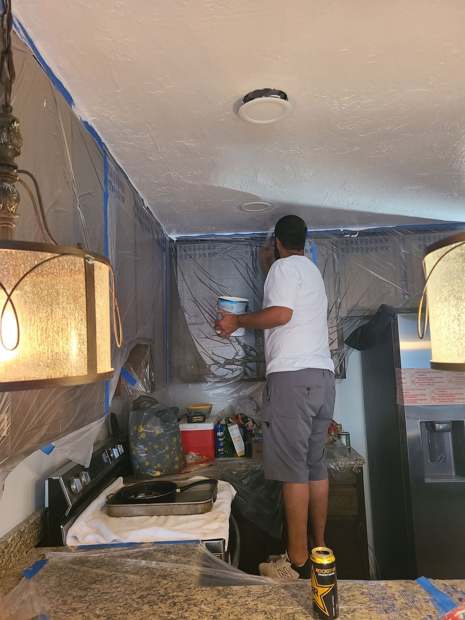 A person in a white shirt and gray shorts stands on a counter to paint a ceiling covered in plastic sheets.