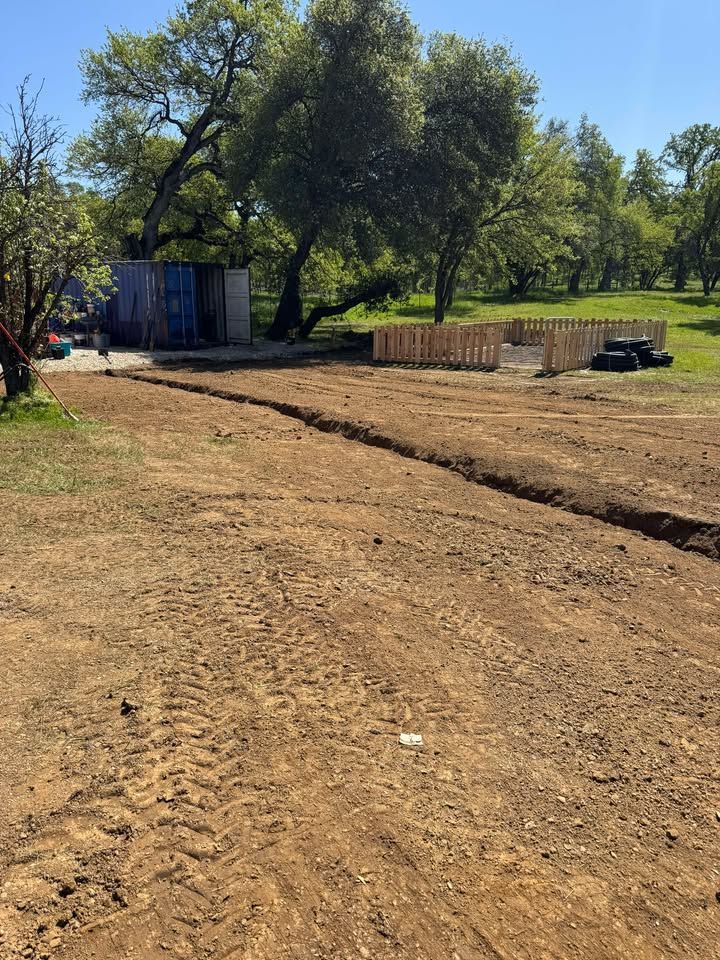Freshly tilled dirt ground in a rural yard with a small wooden fence, a shed, and large trees in the background.