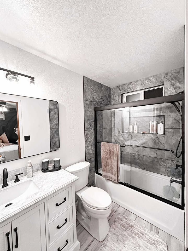 A modern bathroom with a white vanity, a grey marble tile shower with black fixtures, and a white toilet.