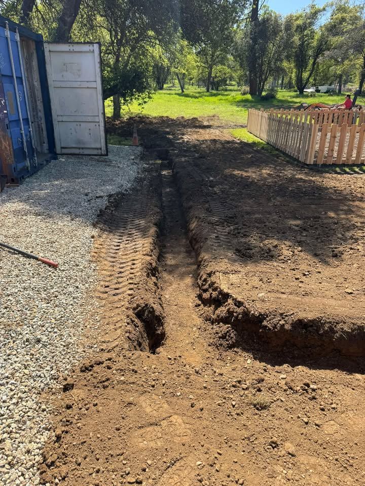 A straight trench dug into bare dirt beside a gravel area and a wooden fence, with trees in the background.