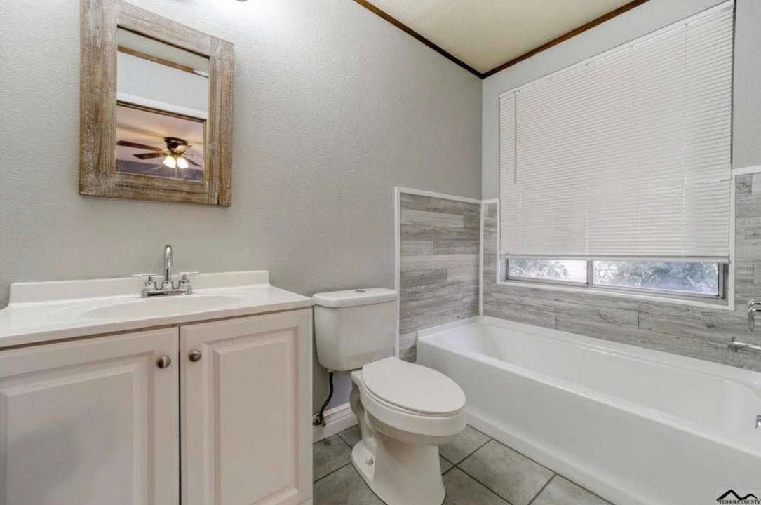 A bathroom featuring a light wood vanity, a mirror, a white toilet, and a bathtub with a window and tiled surround.