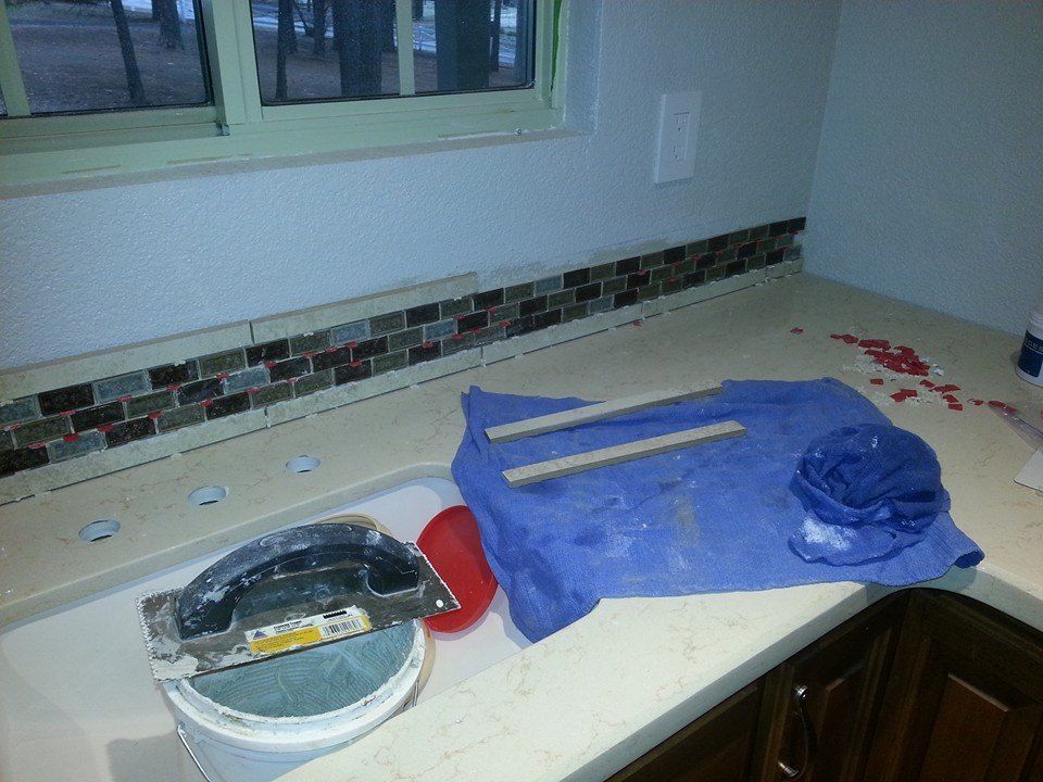A kitchen sink with a grout float in a bucket and a blue towel on the counter, with mosaic tile backsplash in progress.