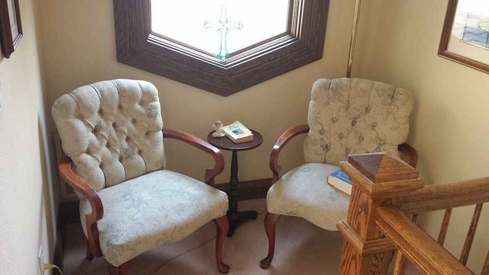 Two tufted armchairs with a small table between them, situated by a corner window with a view of a decorative cross.