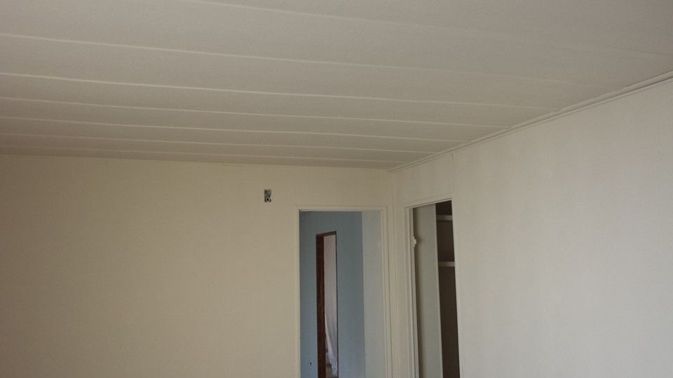 A bright, empty room with plain off-white walls, a paneled ceiling, and a doorway leading to a blue room.