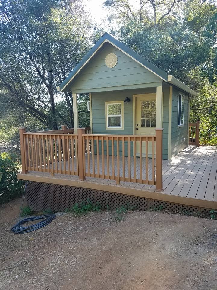 A small, light green cabin with a tan deck and wood railings, set in a wooded area with lattice skirting below the porch.