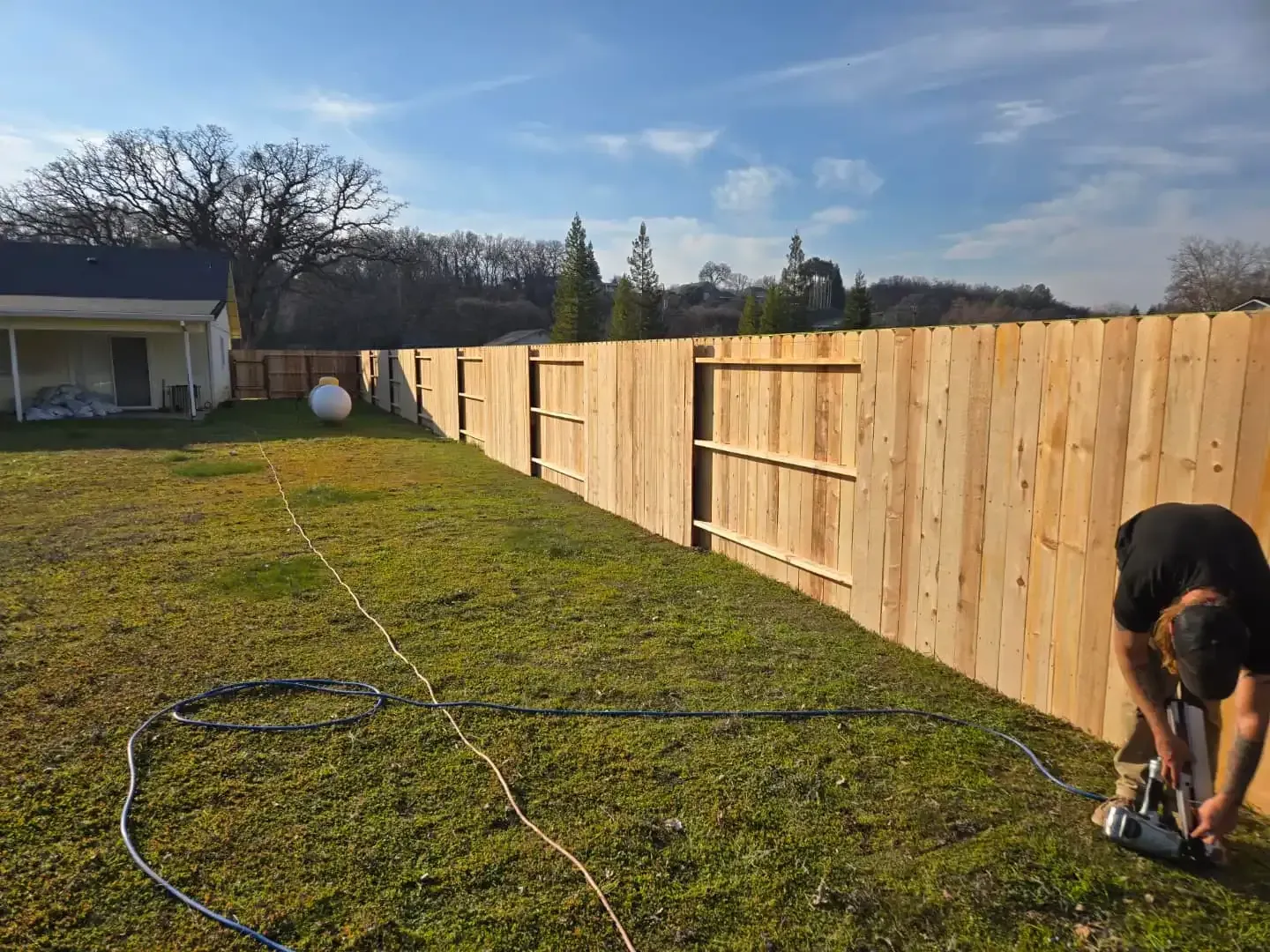A person uses a nail gun to construct a long, unfinished wooden fence in a grassy yard under a bright, sunny sky.