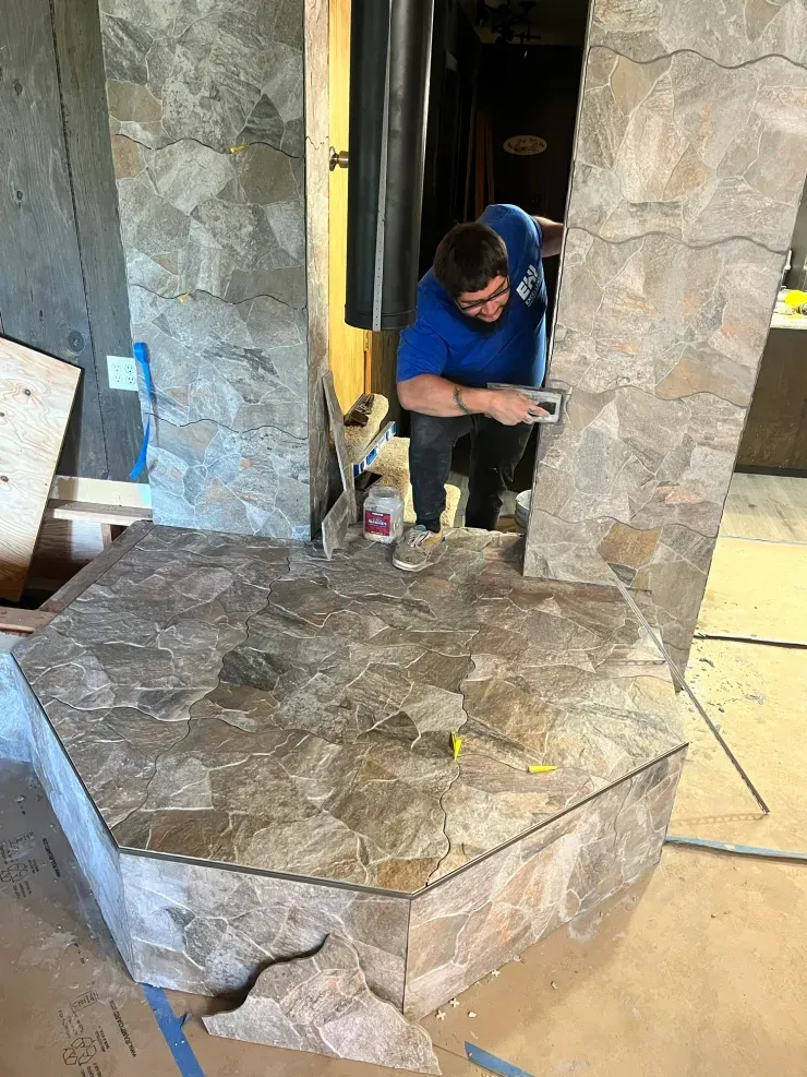 A person in a blue shirt trowels mortar onto a stone-tiled fireplace surround in a room under construction.