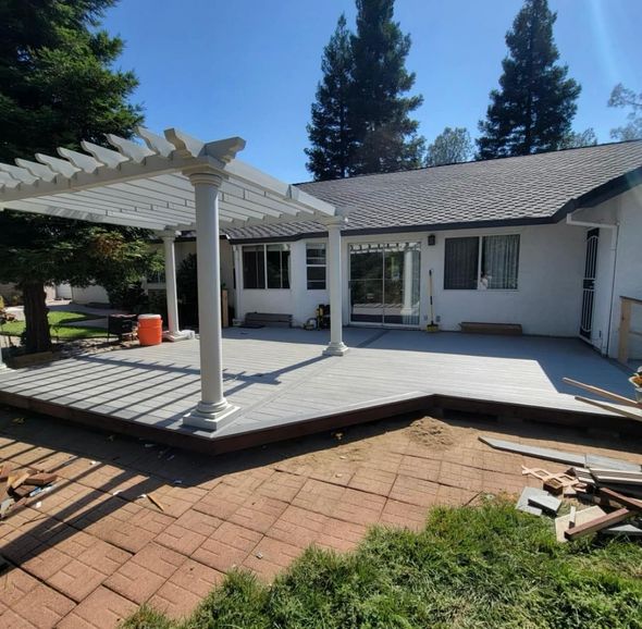 A white pergola stands over a gray deck attached to the back of a house with a shingled roof and red brick patio.