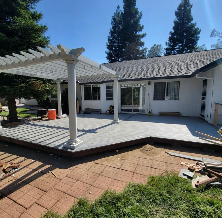 A modern grey deck with a white pergola attached to the back of a light-colored house, next to a brick patio.
