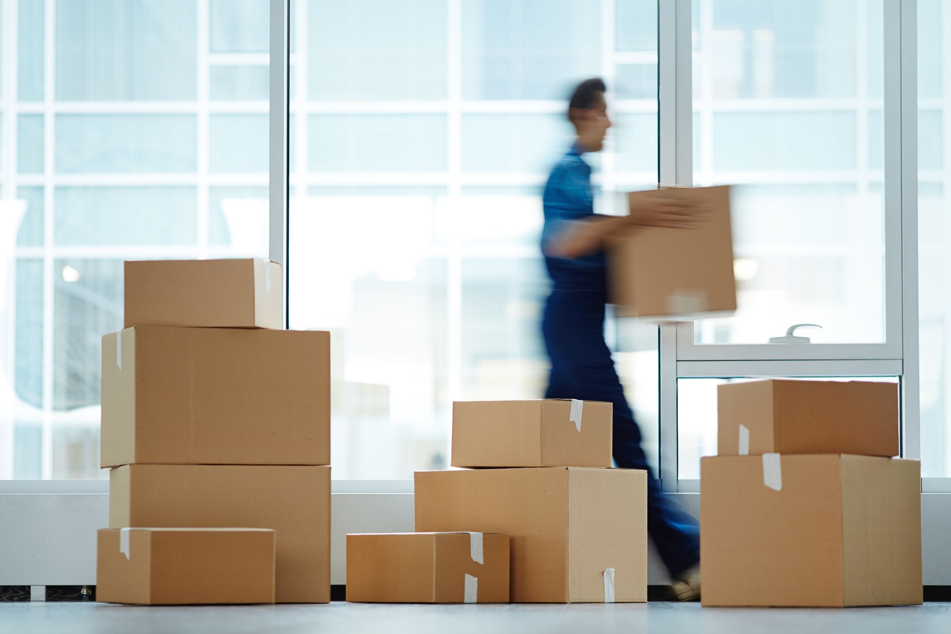 Person carrying a cardboard box among stacked moving boxes in a bright office or warehouse.