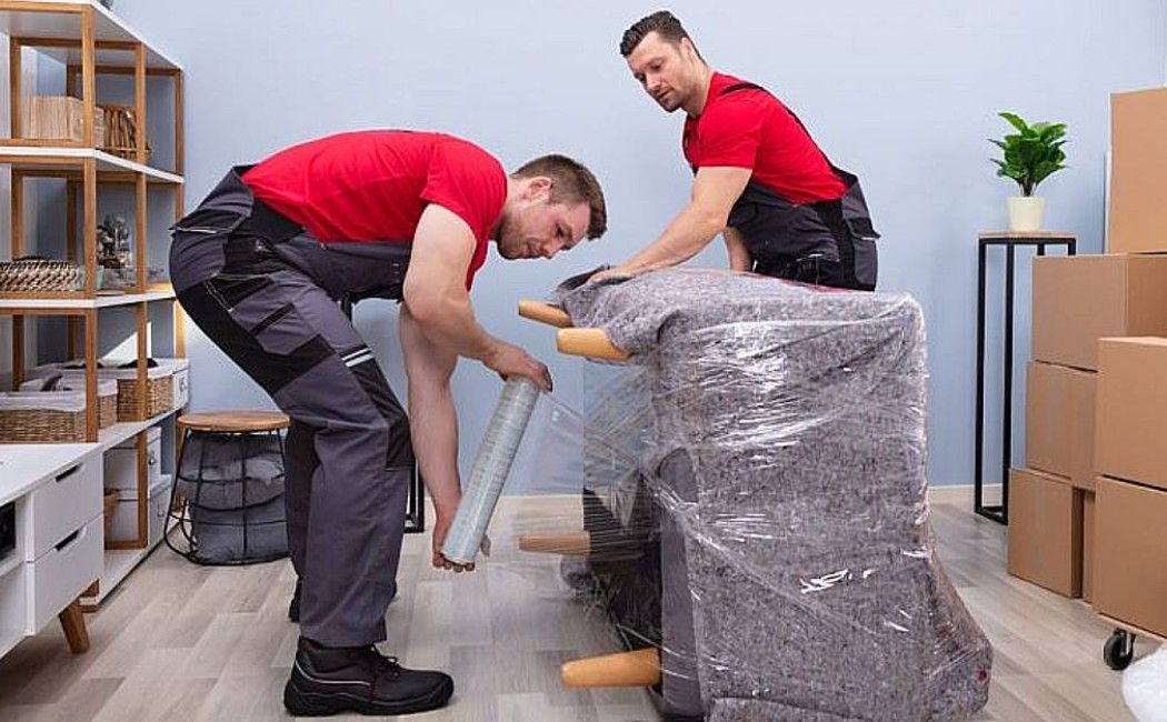 Two movers in red shirts carrying a wrapped chair in a room with boxes and shelving.