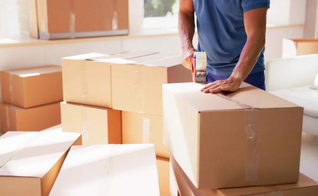 Person packing cardboard boxes in a bright room, moving a large box with tape nearby.
