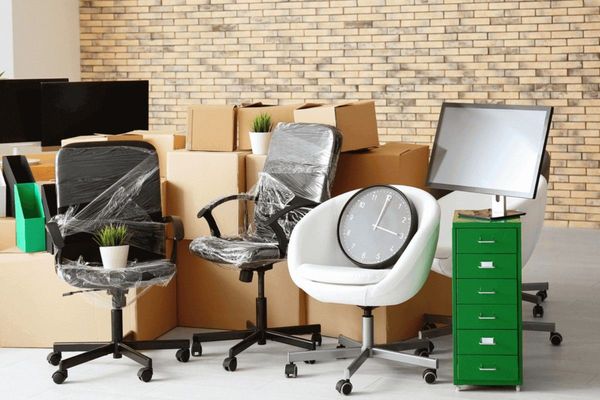 Office chairs and desks arranged in a storage area, with boxes and a green filing cabinet against a brick wall.