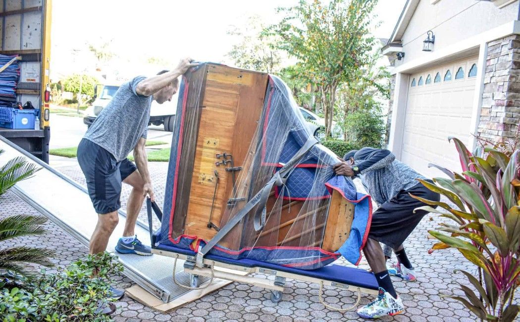 Two people moving an upright piano on a dolly outside a house with blue straps