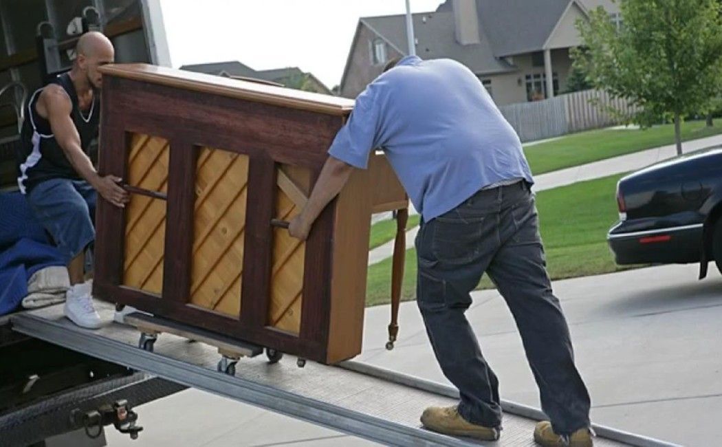 Two men pushing a piano up a ramp onto a moving truck in a suburban driveway