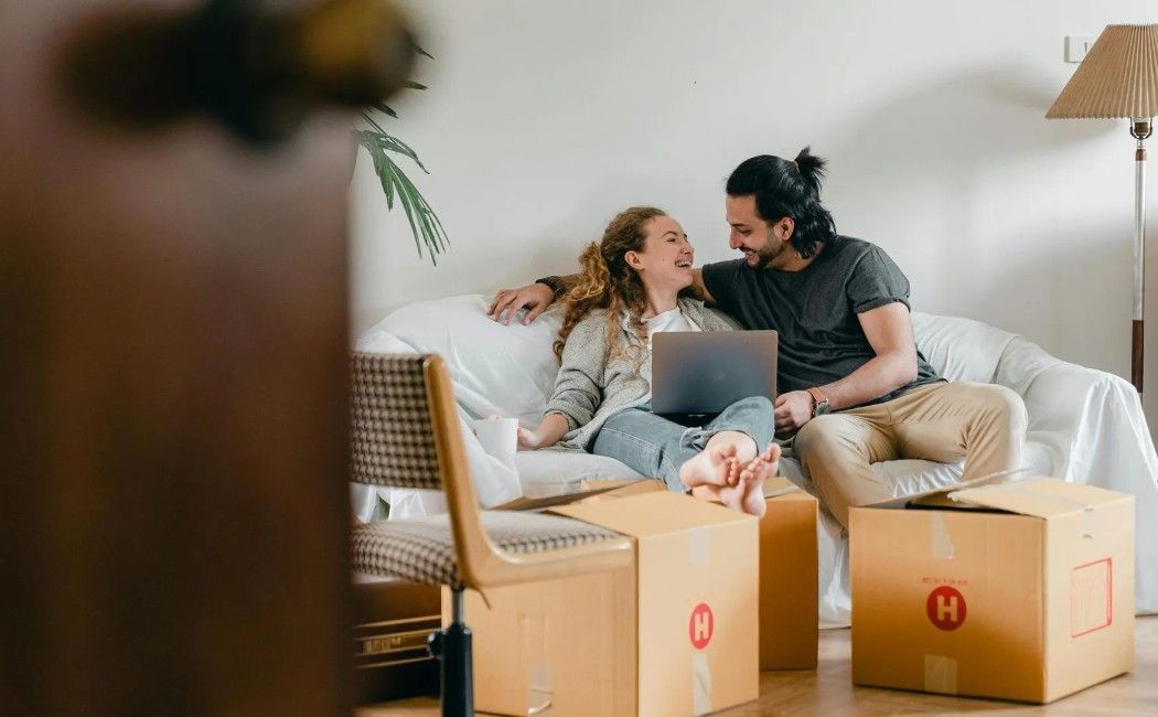 Couple sitting on a couch among moving boxes, smiling at a laptop in a bright living room.