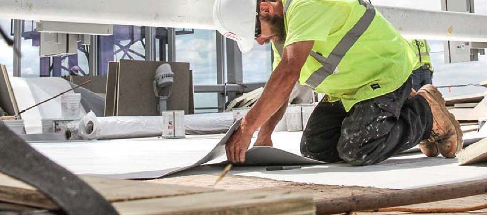 A construction worker is kneeling down on the ground while working on a roof.