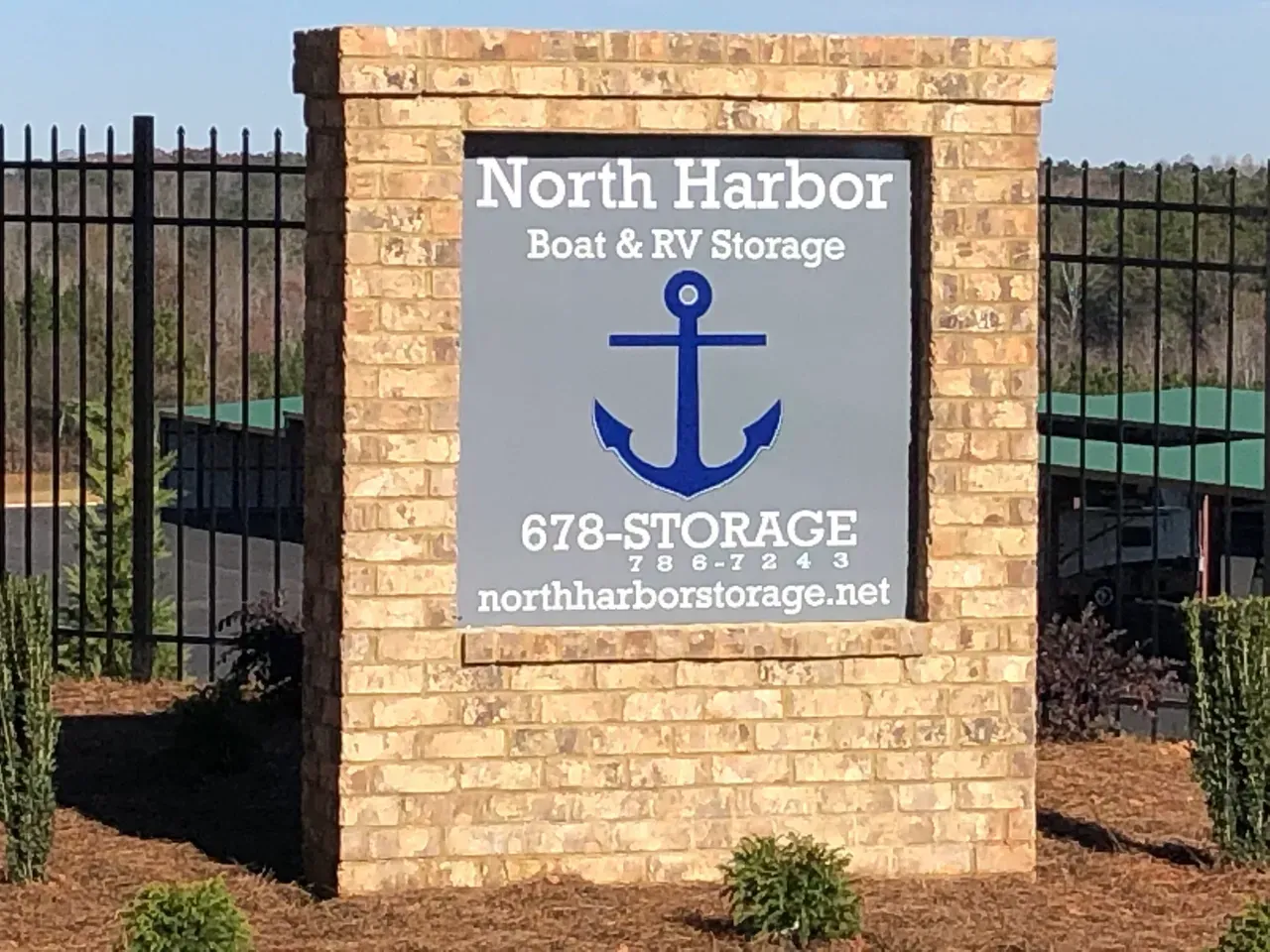 Sign for North Harbor Boat & RV Storage, blue anchor on gray background, brick pillars, black fence.