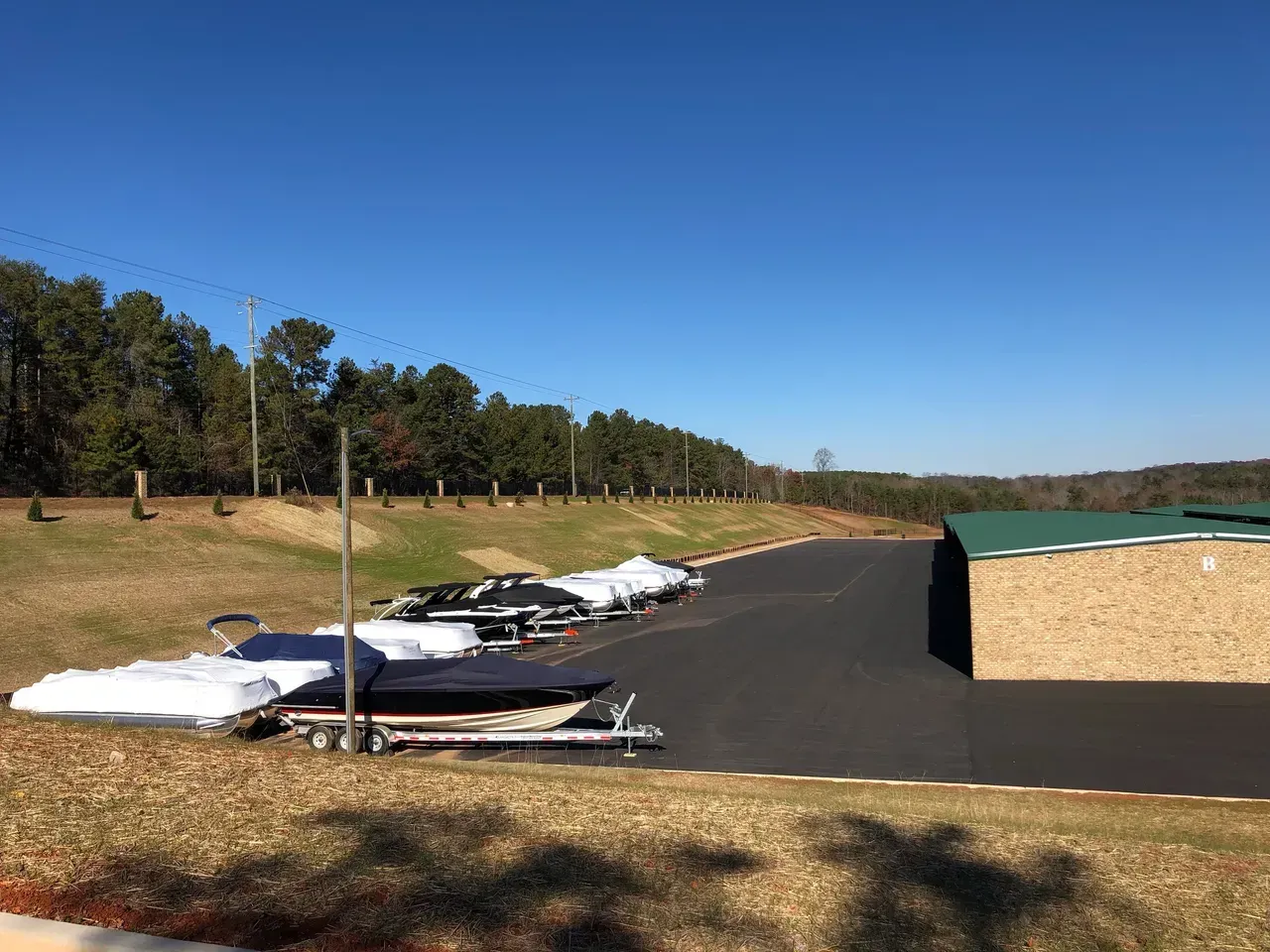 Boats on trailers parked on asphalt, next to a building and hill, under a clear blue sky.
