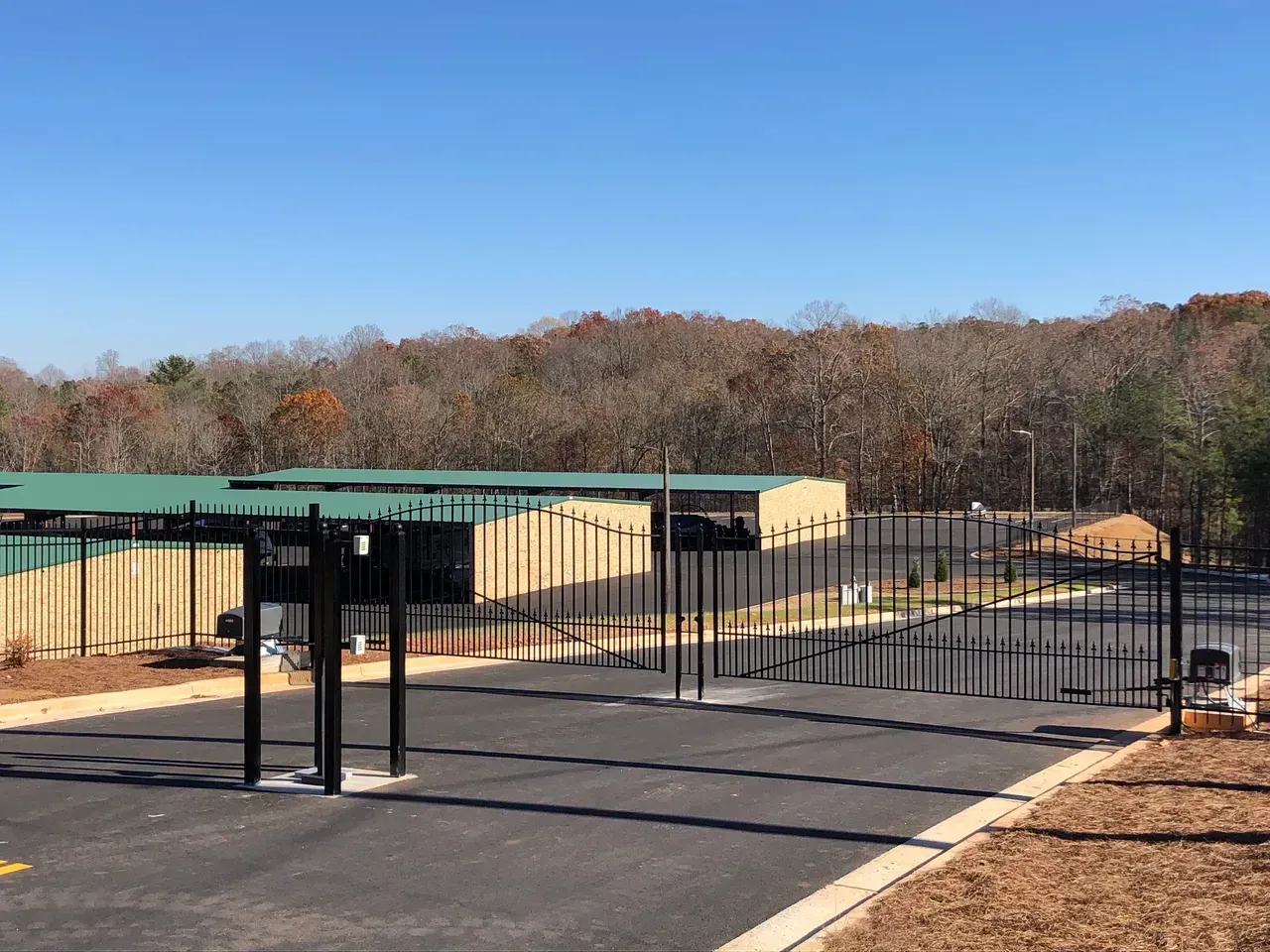Black gated storage facility entrance with tan units, green roofs, and trees against a blue sky.