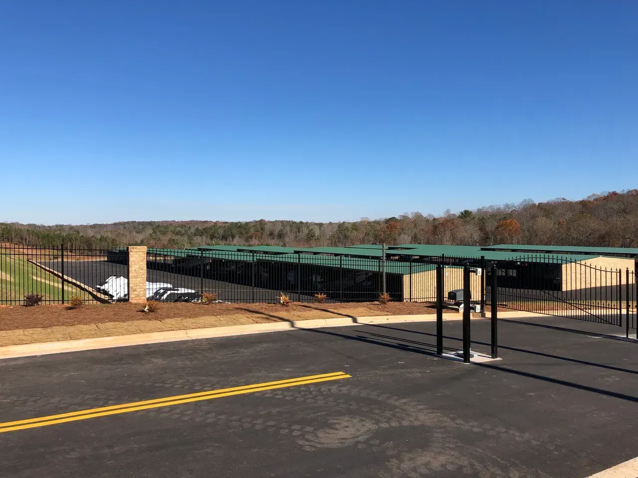 Black fence, buildings, and green-roofed shelters along a road against a treed horizon under a clear blue sky.