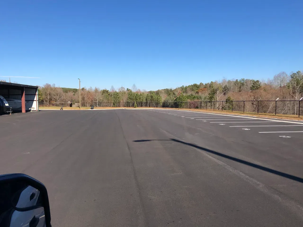 Paved parking lot with a clear blue sky, lined with trees and a building.
