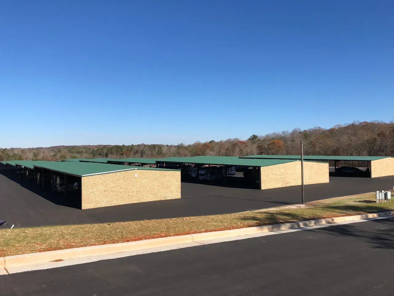 Carports with tan brick walls and green roofs on a black asphalt lot, with a forest background under a blue sky.