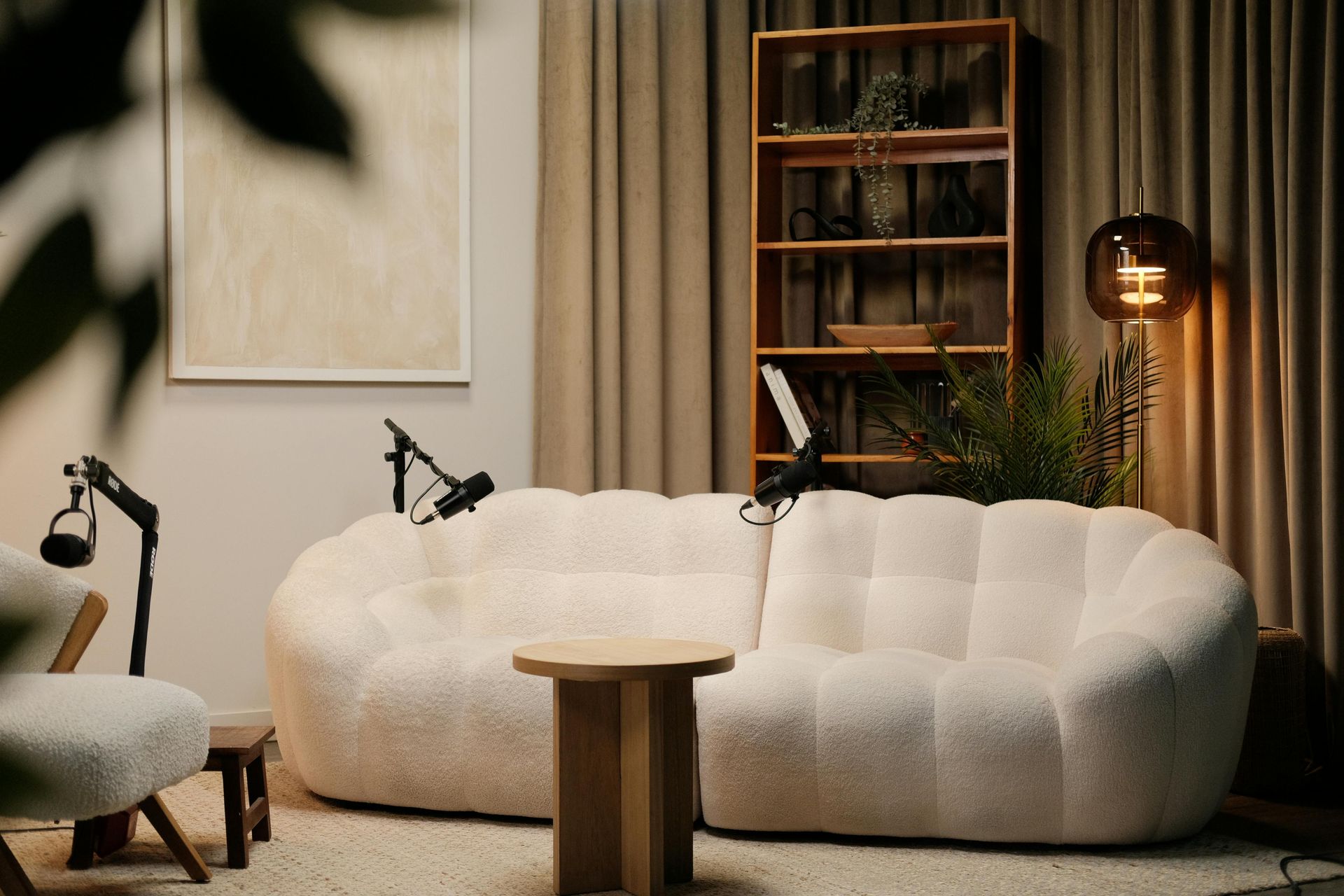 Podcast studio with a white, puffy couch, microphones, and a bookshelf in a warm-toned room.