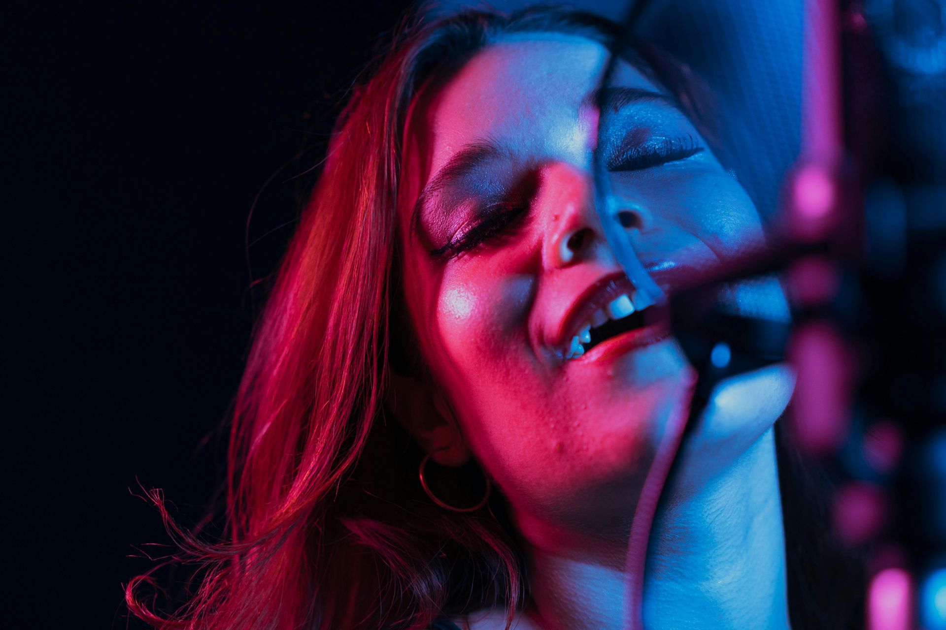 Woman singing into a microphone, lit with pink and blue lights, eyes closed.