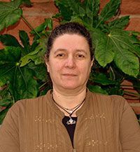 A woman in a brown jacket and necklace is standing in front of a plant.