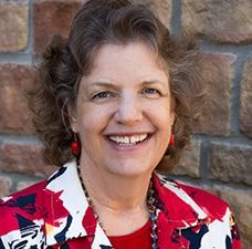 A woman in a red and white jacket is smiling in front of a brick wall.