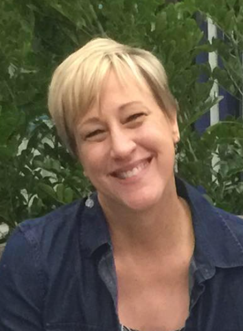 A woman in a blue shirt is smiling in front of a tree.