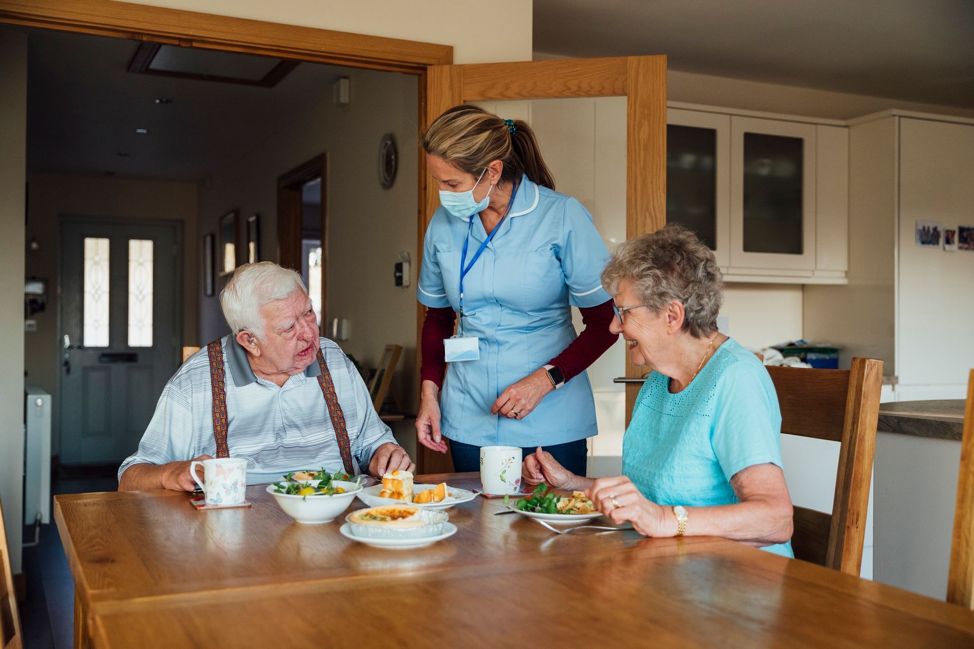A nurse is serving food to two elderly people at a table – North Baldwin, NY - Caring Shepherd HHC