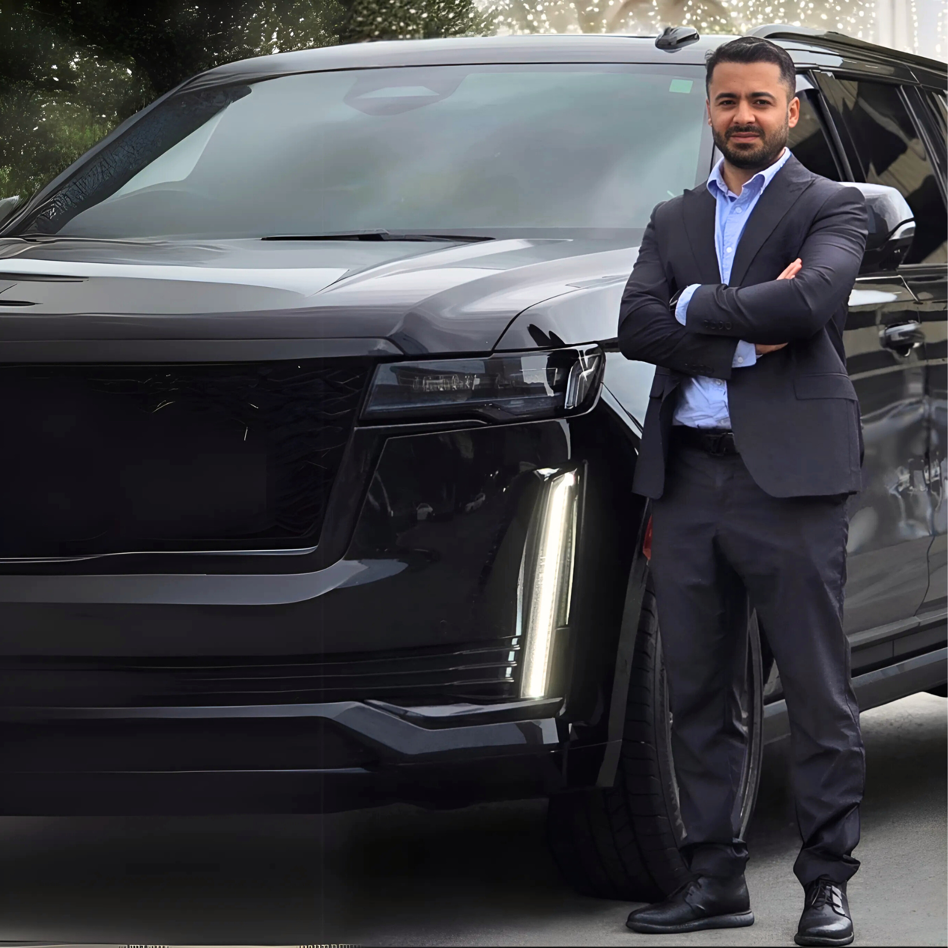 Man in suit standing by a black SUV, arms crossed. Outdoor setting.