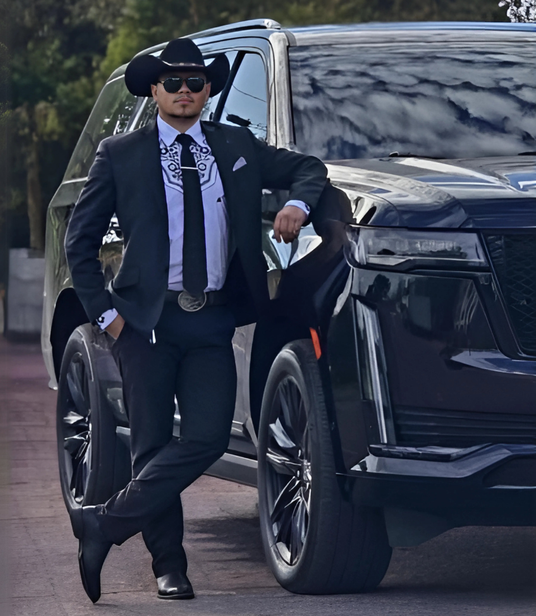 Man in black suit and cowboy hat leans against a black SUV.