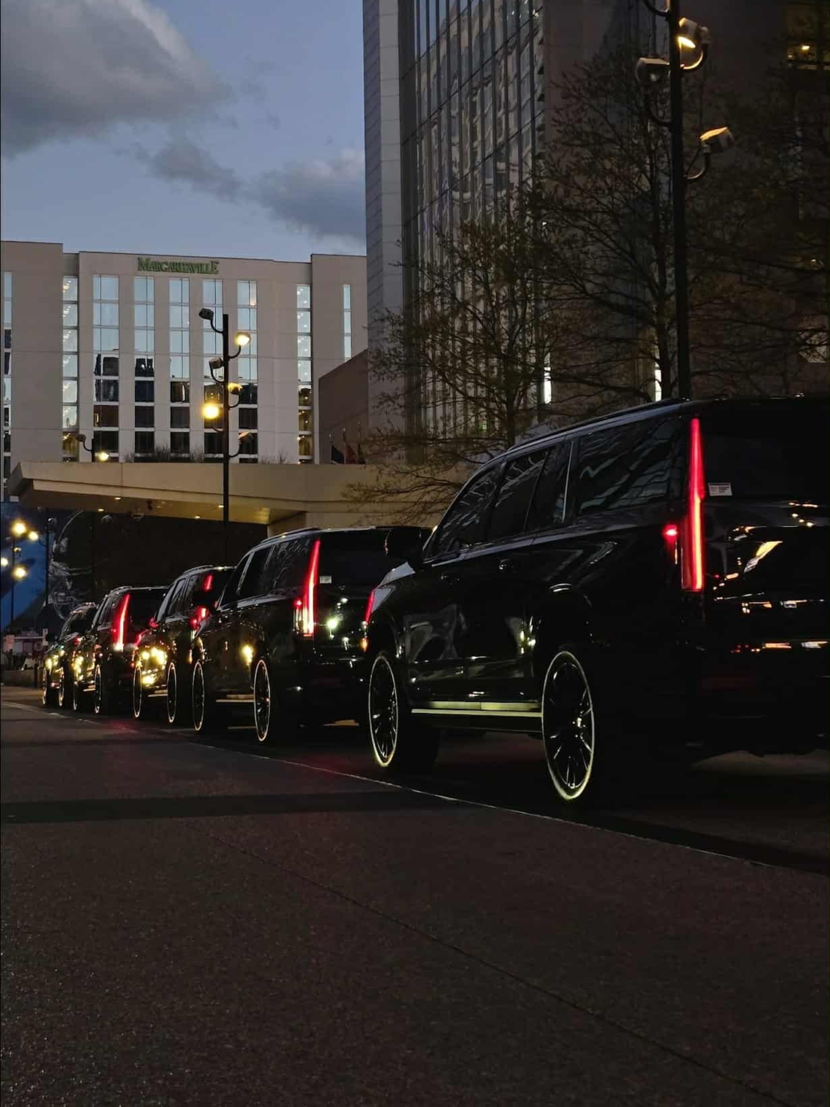 Black SUVs parked on a street in front of a modern building with a clear glass facade. Evening setting.