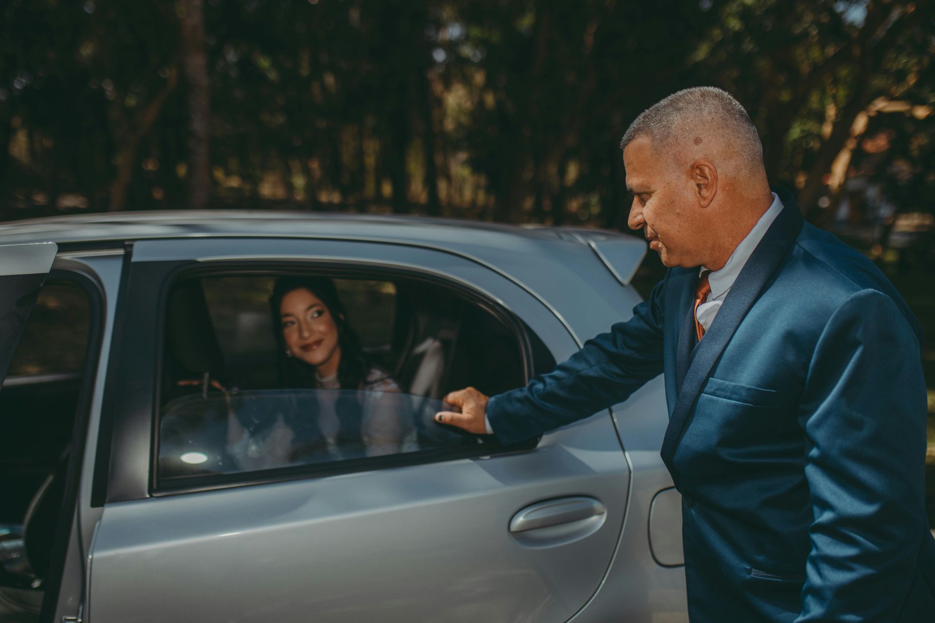 Man in a blue suit opening a car door for a person inside; outdoor setting.