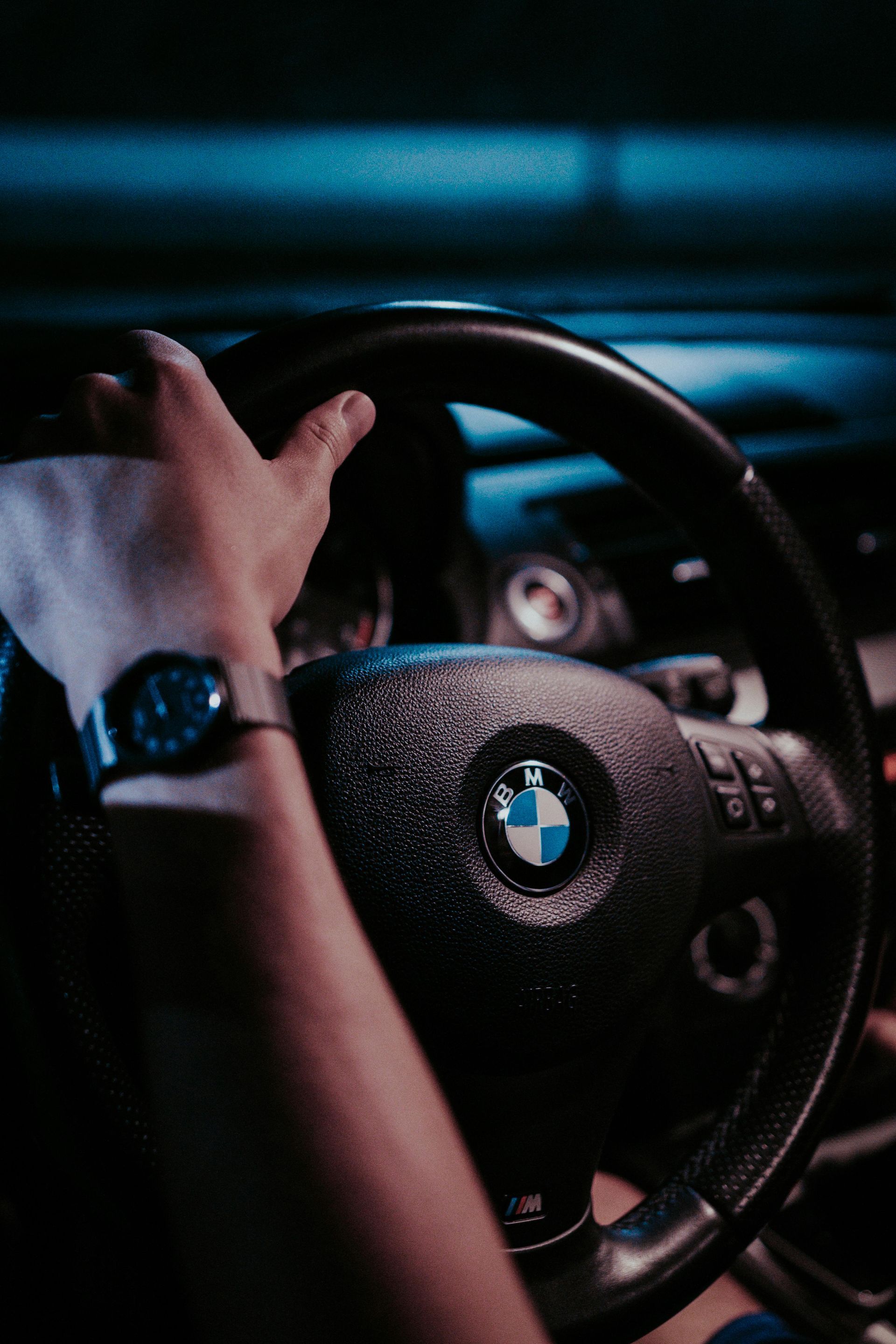 Person's hands gripping a black BMW steering wheel with blue lighting. A wristwatch is visible.