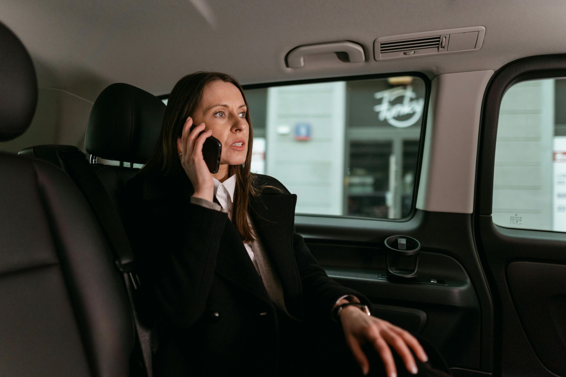 Woman in a car, talking on the phone. She wears a black coat and a white shirt.