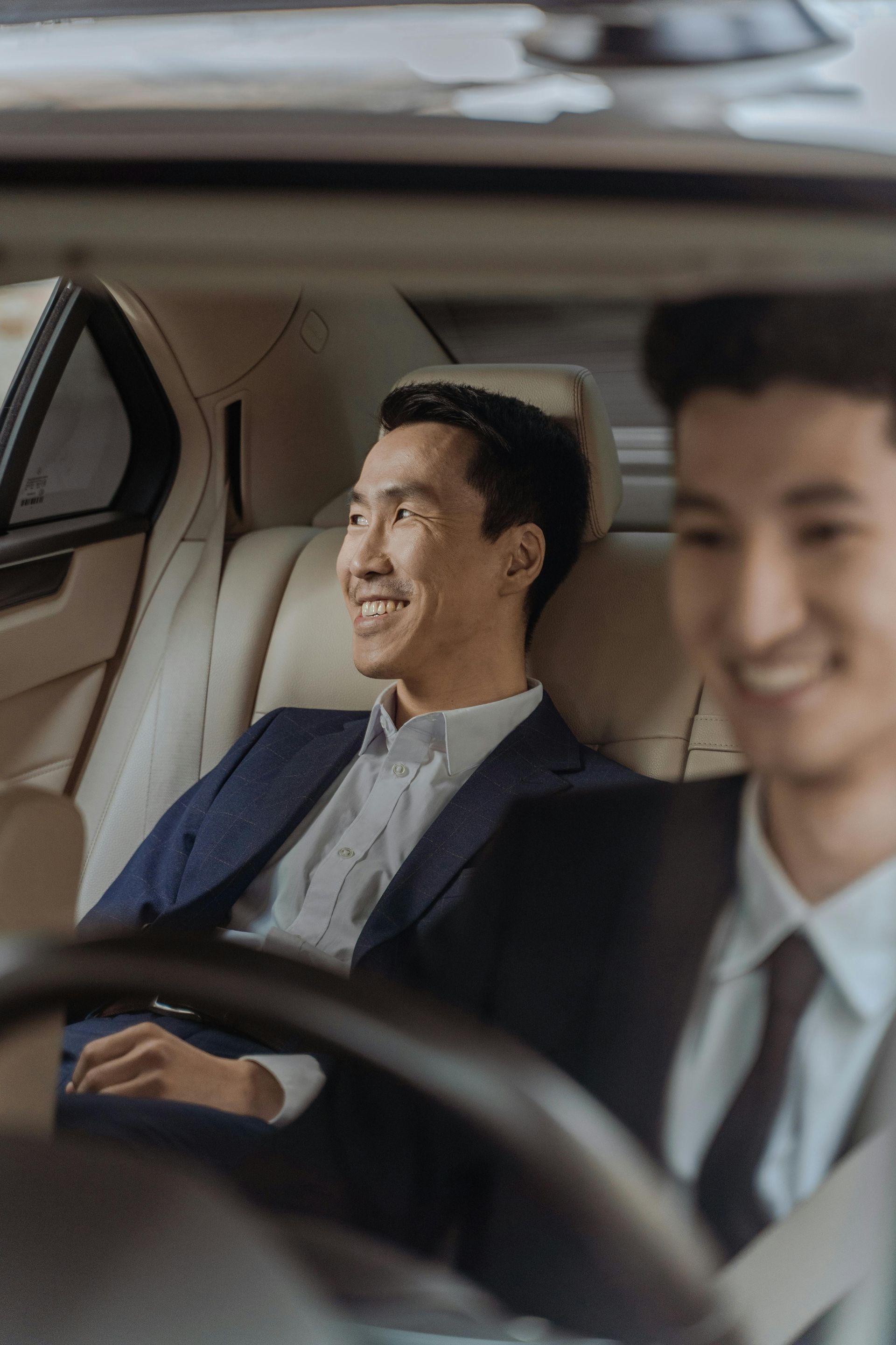Two men in suits smiling inside a car. One is in the backseat.