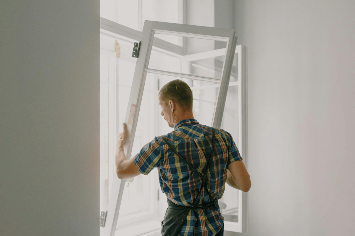 Man installing a window, wearing plaid shirt and apron. Indoors, white walls.