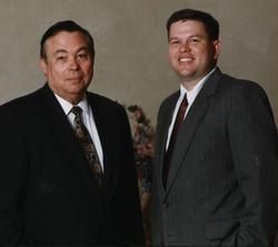 Two men in suits posing for a photo. One smiles; the other looks forward. Beige backdrop.