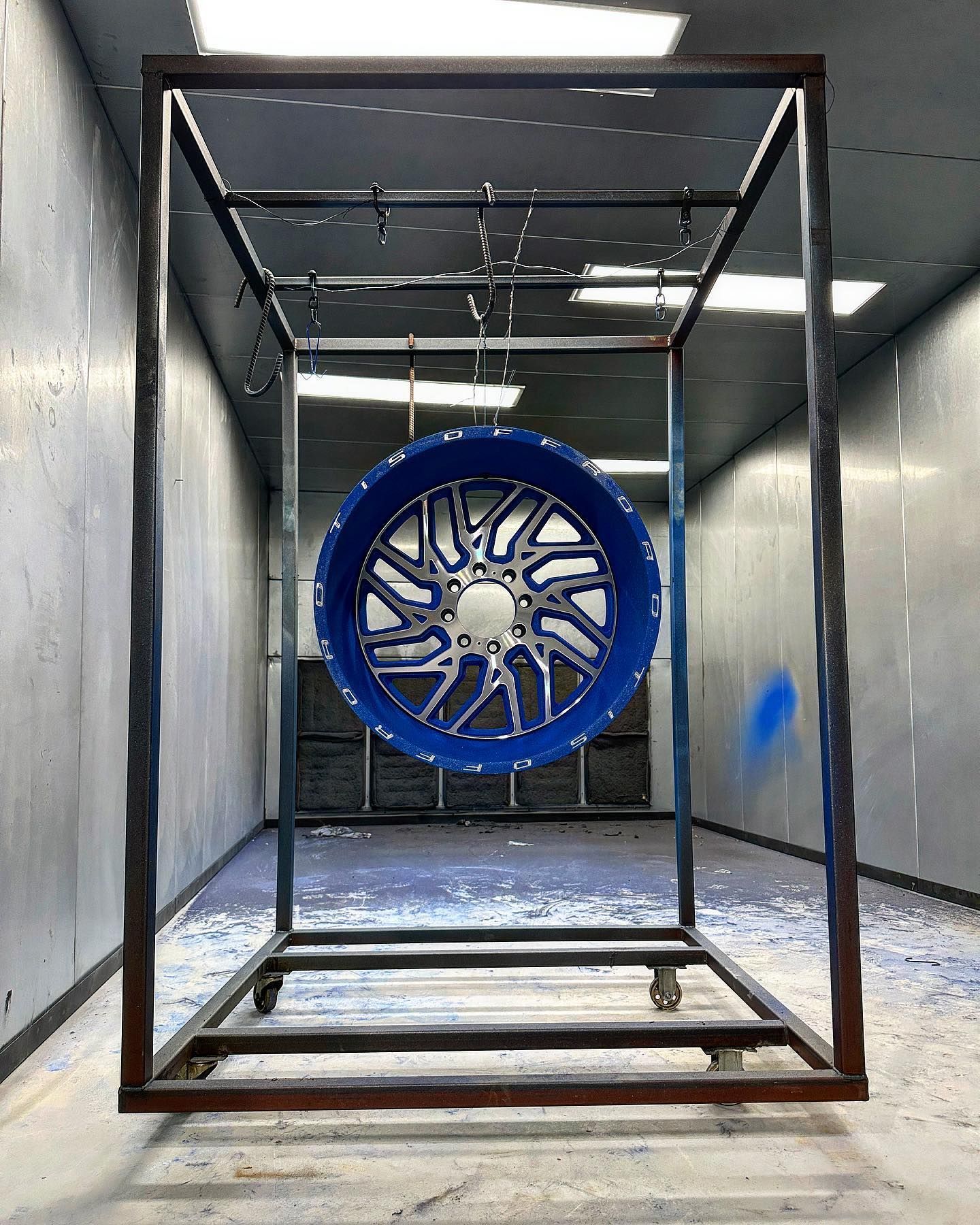 Blue and silver car wheel hanging inside a metal frame in a powder-coating booth.