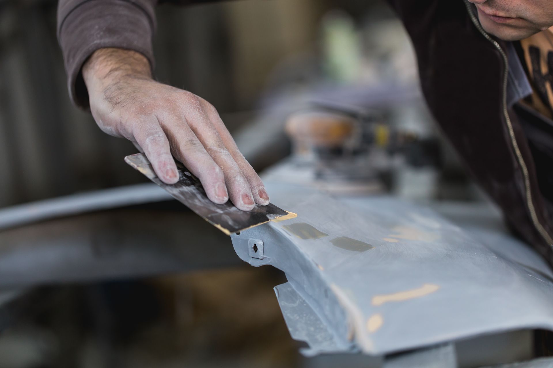 Person sanding a gray car bumper with a metal sanding block in a workshop.